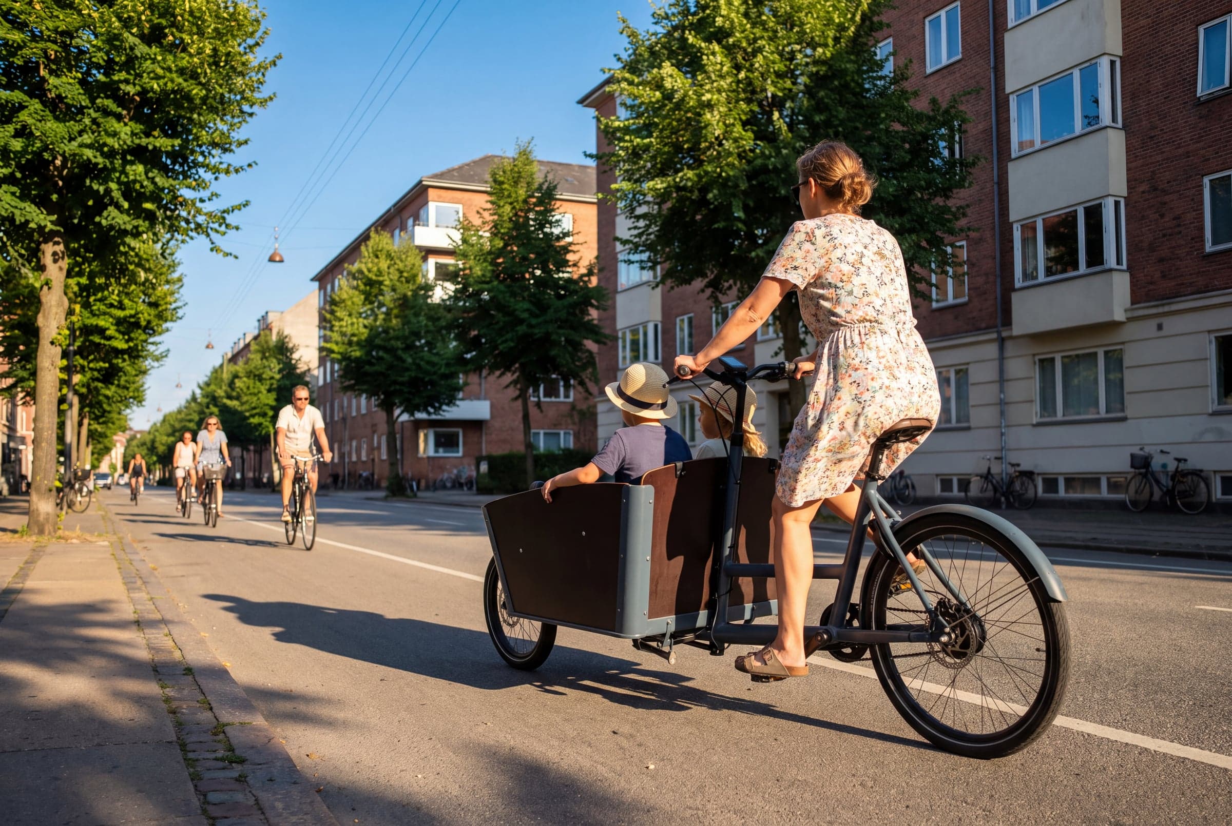 A parent rides a Sykla Cargo bike with two children in the box along a wet Copenhagen cycle lane.